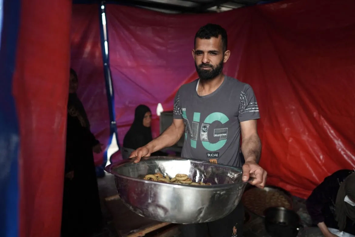 وسطی غزہ کی پٹی دیر البلاح میں عید الفطر کے موقع پر ایک شخص خیمے کے اندر فلسطینی خواتین کی جانب سے بنائی ہوئی کوکیز کا پیالہ اٹھائے ہوئے ہے۔ [تصویر بذریعہ اے ایف پی]